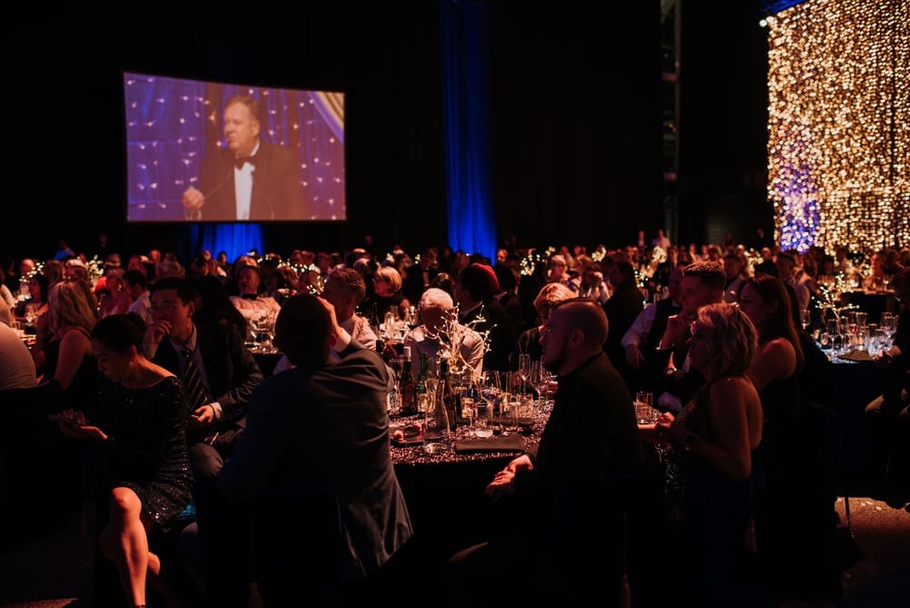 A formal event with many people seated at round tables, watching a man in a tuxedo speaking on stage, shown on a large screen. The room is dimly lit with blue and warm string lights.