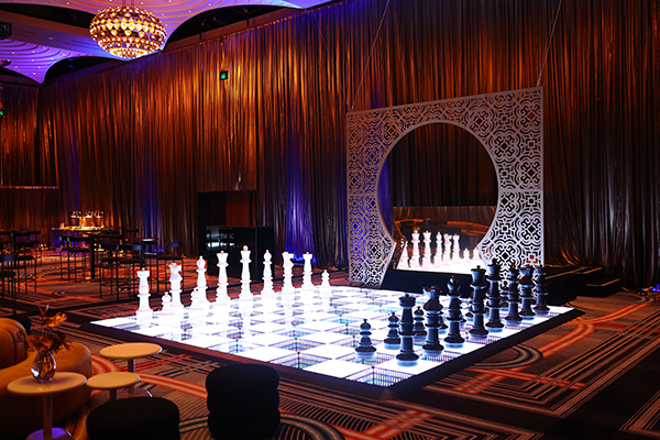 A large, illuminated chessboard with oversized black and white chess pieces is set up in an elegant, dimly lit room with ornate décor and draped curtains, creating a luxurious and dramatic atmosphere.