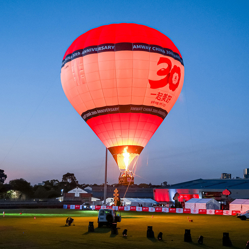 A large red and white hot air balloon with Amway China 30th Anniversary and 30 printed on it is illuminated at dusk on a grassy field, showcasing the expertise of an event production Melbourne team, with buildings and tents in the background.