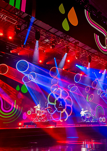 A vibrant stage with colorful neon lights and cutting-edge event technology, abstract shapes and patterns projected on a large screen, featuring two musicians playing drums under red, blue, and purple lighting effects.