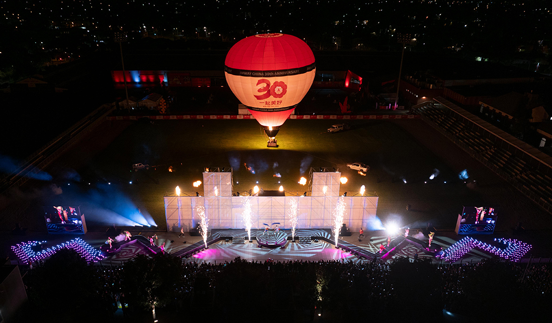 Aerial view of a nighttime event in a stadium with a hot air balloon marked 30 floating above a stage, bright lights, fire effects, and a large audience seated on either side.
