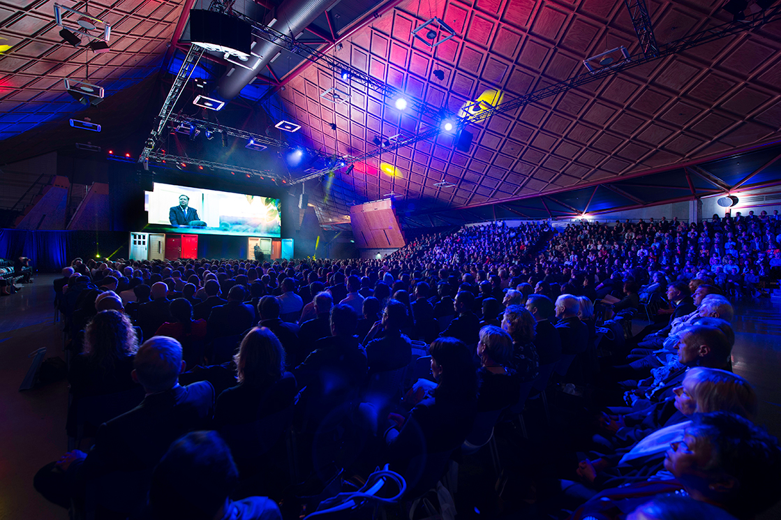 A large audience sits in a spacious, modern auditorium with a geometric ceiling, watching a brightly lit stage where the conference theme is displayed on a large screen. Blue and purple lights illuminate the space.