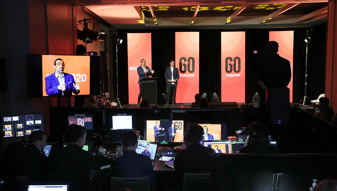 A stage event is being filmed with several people working at computer stations in the foreground. Two speakers stand on stage in front of tall orange “GO Foundation” banners, and a large screen displays a man speaking.