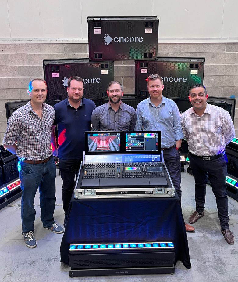 Five men stand indoors behind a lighting control console and screens, with large Encore-branded equipment cases stacked behind them and LED lights on the floor, in a professional setting.