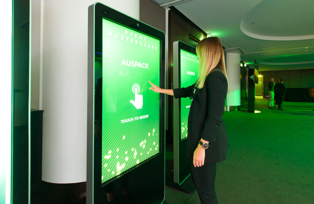 A person in business attire touches a large interactive screen that displays EVENT MASTERCLASS and AUSPACK in a green-lit modern event space.