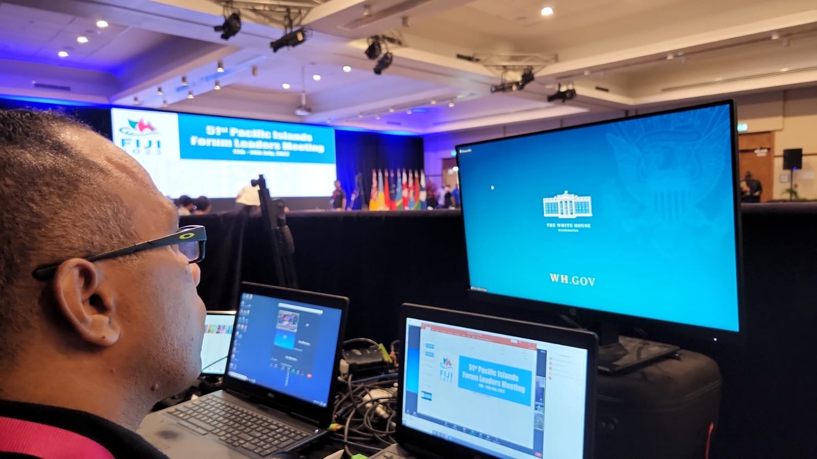 A man sits at a desk with multiple laptops and monitors provided by an event production company Wollongong at the 51st Pacific Islands Forum Leaders Meeting. The White House logo is on screen, with a stage and flags in the background.