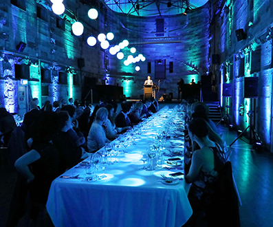 A long dining table set for a formal event is surrounded by guests in a grand, stone-walled hall with blue lighting and glowing orbs overhead, enhanced by cutting-edge event technology. A speaker stands at a podium at the end of the table.