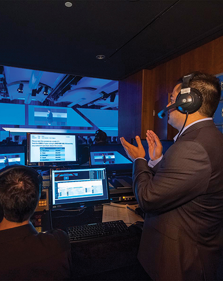 Two people wearing headsets work at a control booth with multiple monitors, managing an event with a blue-lit stage visible through a window. One person is clapping, perhaps celebrating an encore performance.