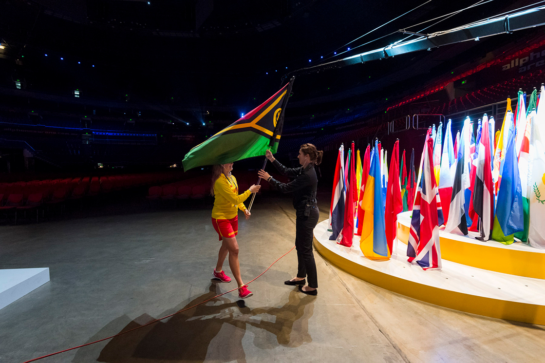 A person hands a large Vanuatu flag to another individual near a display of various national flags on a circular platform in an indoor stadium with dim lighting.