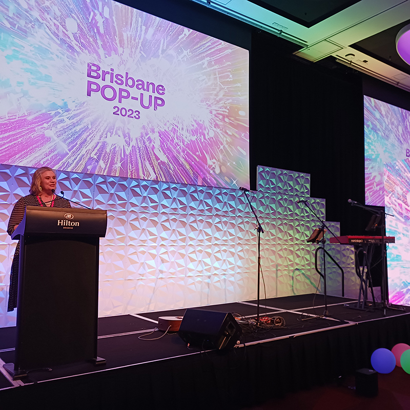 A woman stands at a podium on stage with a sign reading Brisbane POP-UP 2023 displayed behind her. The venue, featuring Hilton branding and stage equipment, showcases the expertise of an events AV company Brisbane trusts.