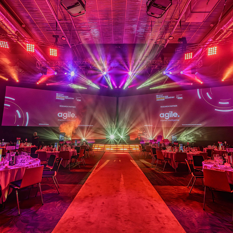 A large event hall with round tables set for dinner, dramatic stage lighting in pink and purple hues, and a stage displaying agile. on big screens—expertly styled by Encore Canberra, your trusted event production Canberra team.