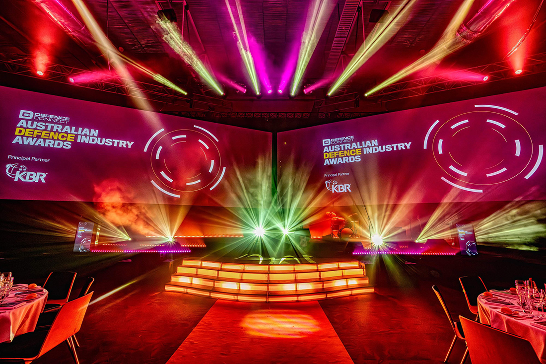Colorful stage set for gala dinner with dramatic lighting and big screens displaying “Australian Defence Industry Awards.” Round tables fill the venue as vibrant beams of pink, orange, and green lights highlight the stage.