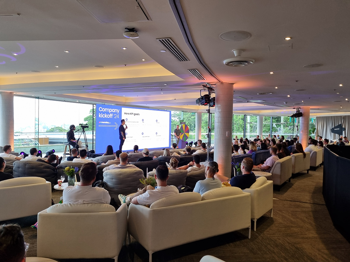 A large group sits on white couches in a spacious, modern room, watching a presenter speak at Company Kickoff 24 with slides projected behind them. Floor-to-ceiling windows reveal greenery outside—an event expertly produced by Encore Sydney.