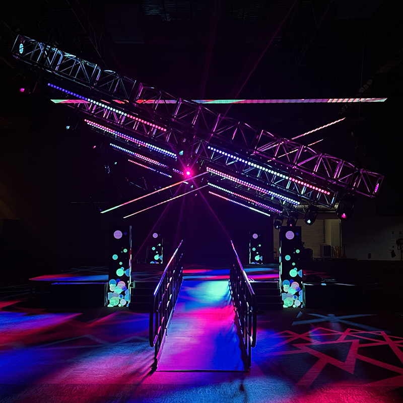 A stage with a ramp and railings is illuminated by colorful spotlights and LED lights arranged on overhead trusses, creating a vibrant, futuristic atmosphere in a dark indoor space.