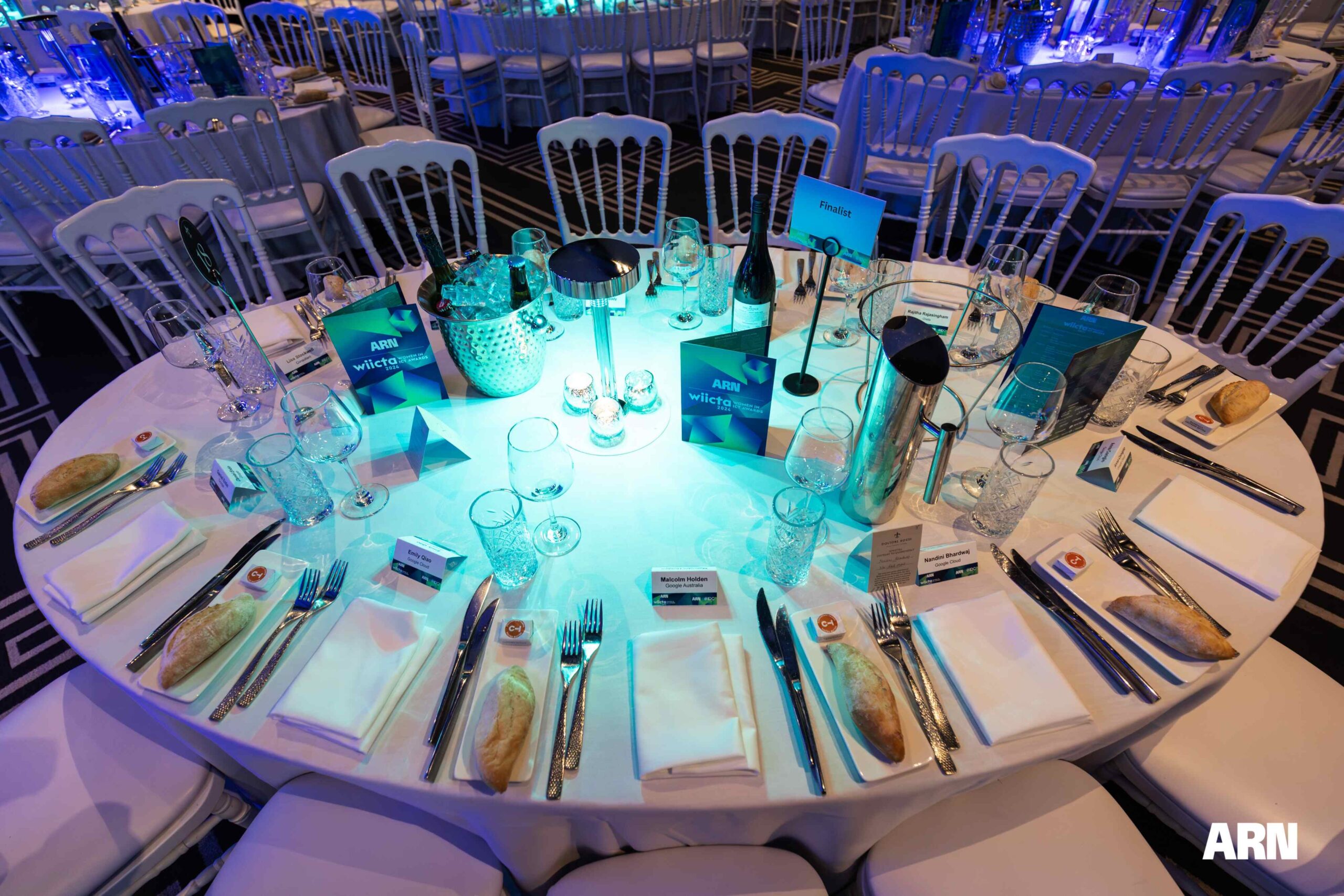 A round banquet table set for a formal event with plates, cutlery, glasses, bread rolls, menus, and name cards under blue lighting. Elegant white chairs surround the table. The ARN logo is visible in the corner.