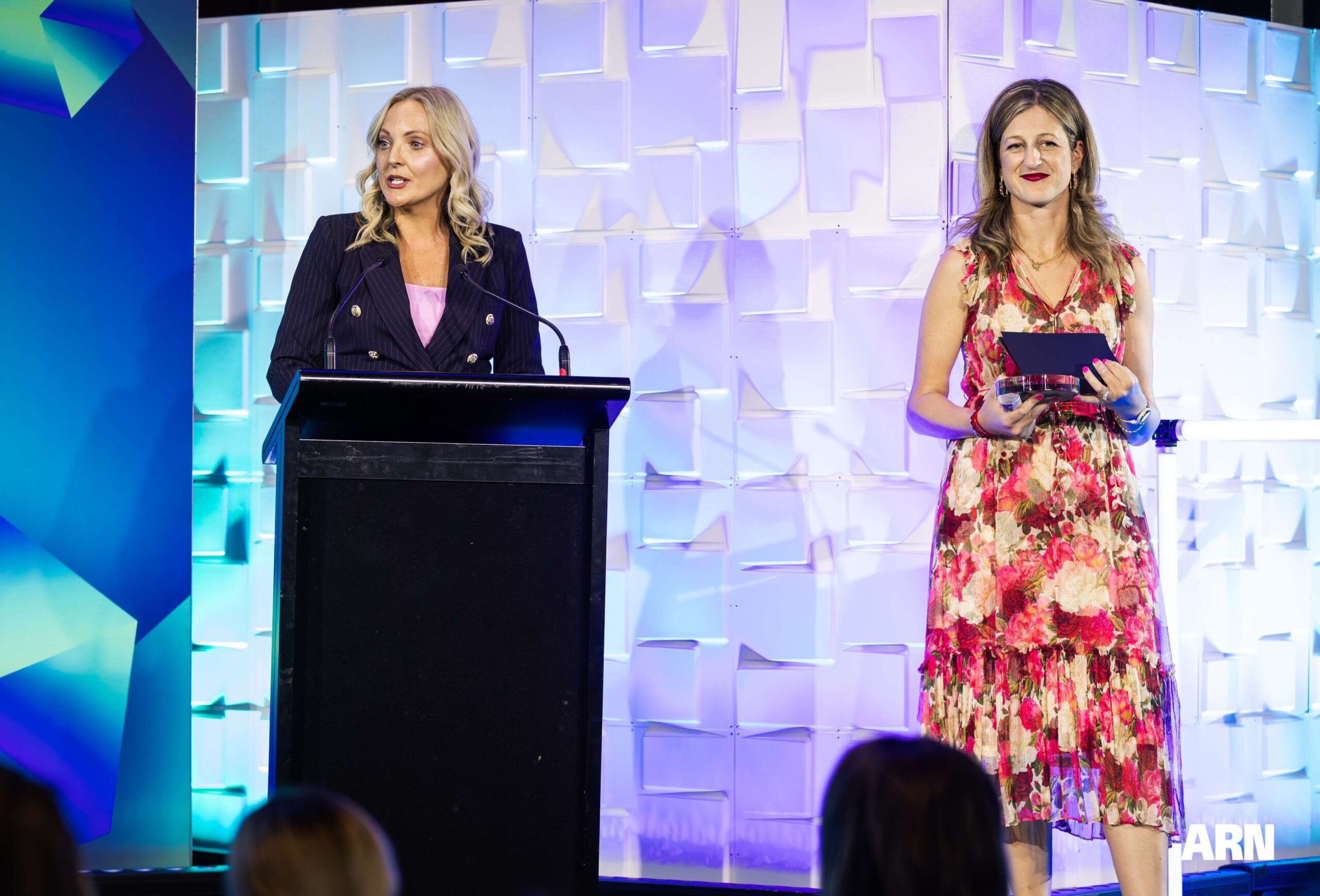 Two women on stage at an event; one stands at a podium speaking, wearing a dark blazer, and the other in a floral dress holds a tablet. The background features geometric patterns and colorful lighting.