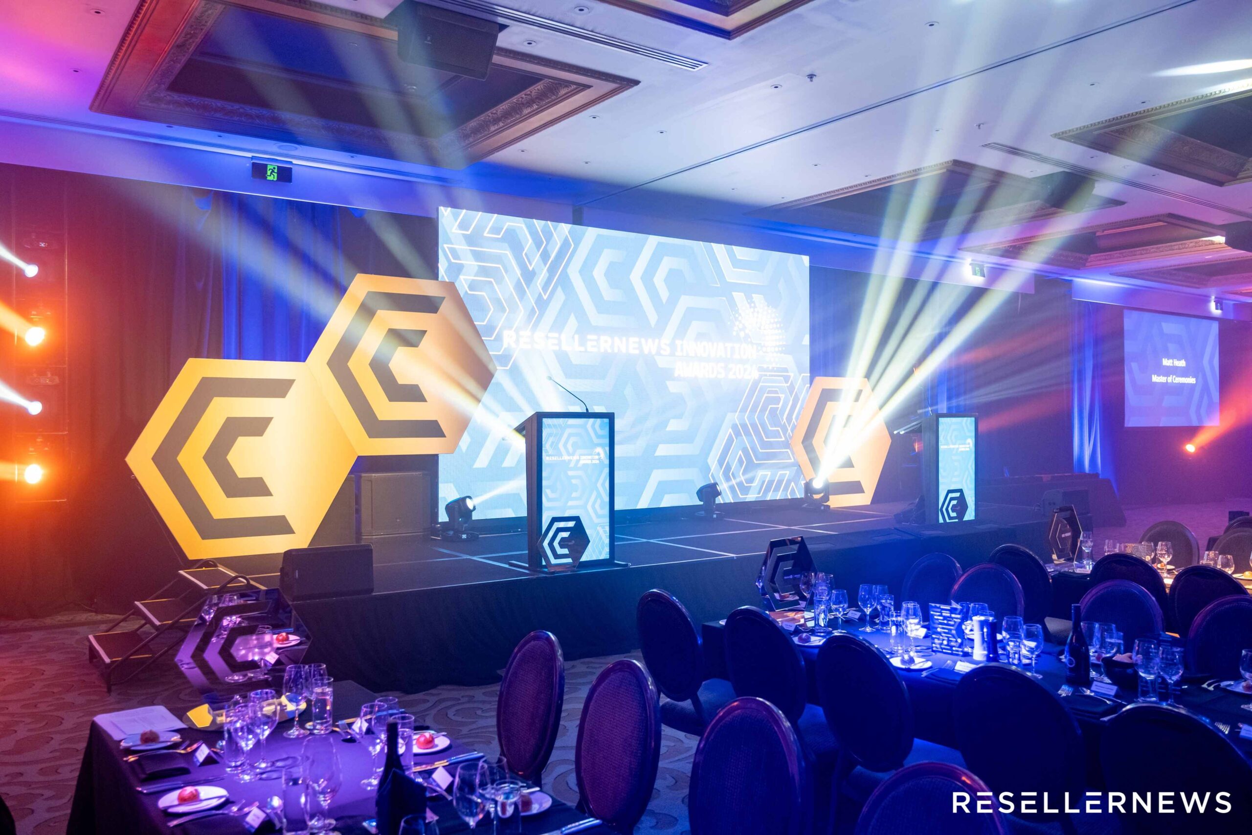 A large, elegant conference room set up for the Reseller News event, featuring a brightly lit stage with geometric designs, podiums, spotlights, and decorated dining tables in the foreground.