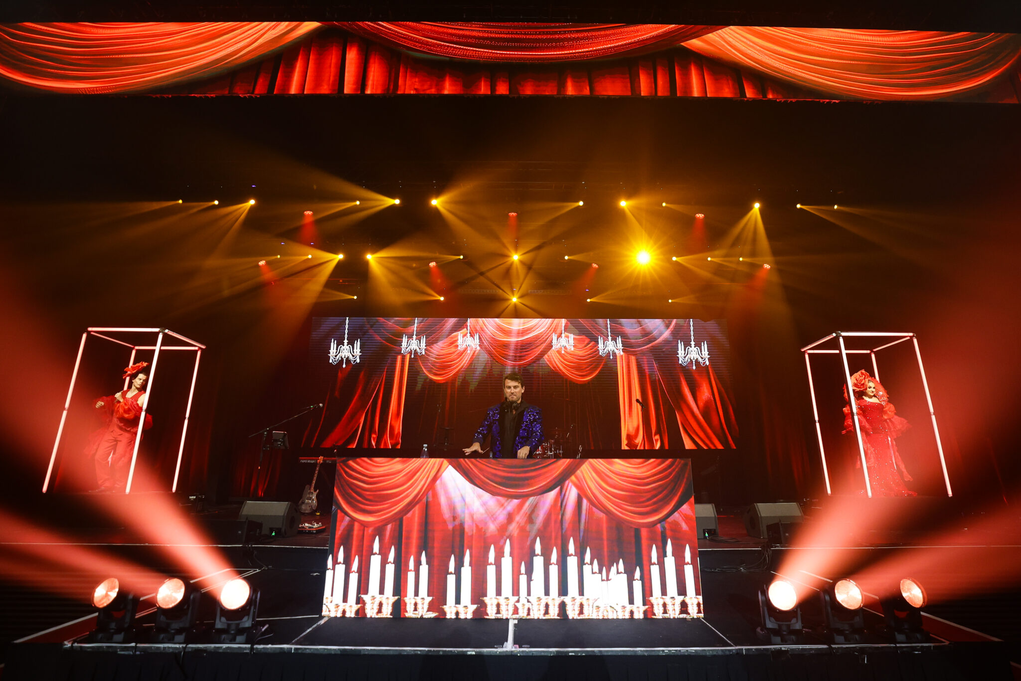A DJ performs on stage with red curtains and chandeliers displayed on screens behind him. Bright yellow and red stage lights shine, with two dancers in illuminated cages on either side of the DJ booth decorated with candle graphics.