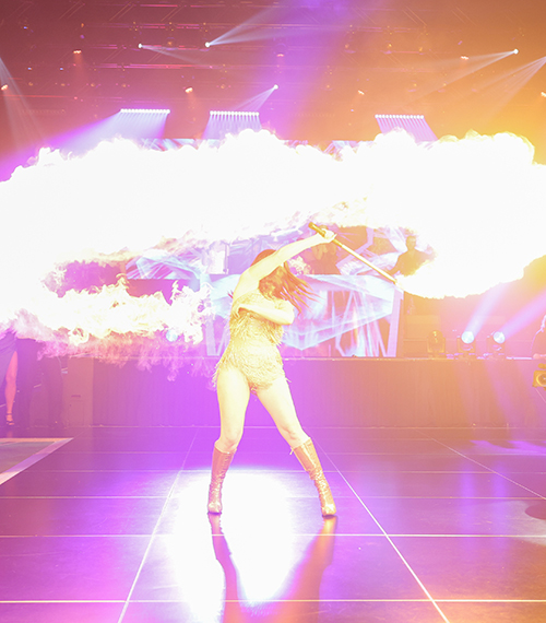 A performer in a sparkling outfit spins a flaming staff on stage, creating a dramatic arc of fire, with bright lights and a DJ booth in the background.