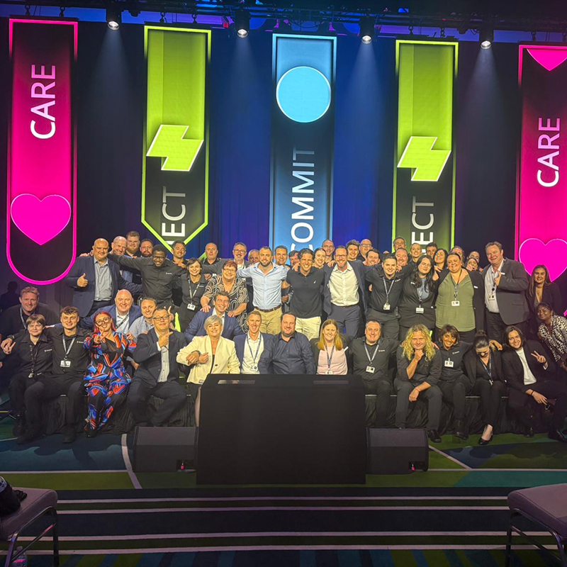 A large group of people pose together on a stage, smiling at the camera. Behind them are colorful banners with the words CARE, COMMIT, and RESPECT, and matching symbols, all under bright stage lighting.