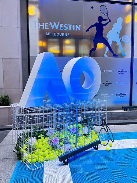 A large AO sculpture stands atop a wire box filled with tennis balls and flowers, with tennis rackets leaning against it. In the background is The Westin Melbourne sign and silhouettes of two tennis players.