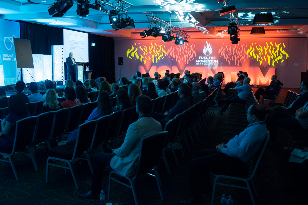 A speaker presents to an audience seated in a dimly lit conference room, with orange lighting and Fuel the Momentum displayed on the back wall—expertly staged by Encore Gold Coast.
