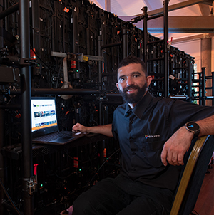 A man with a beard sits at a laptop in front of large racks of electronic equipment, smiling at the camera. He wears a black shirt and a smartwatch. The setting appears to be a technical or server room.
