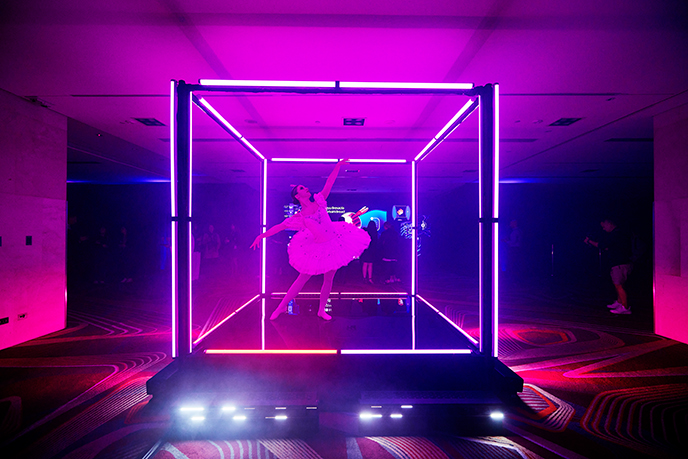A ballerina in a tutu poses gracefully inside a glowing neon cube installation, enhanced by vibrant pink and purple event technology lighting in a dark, modern room.