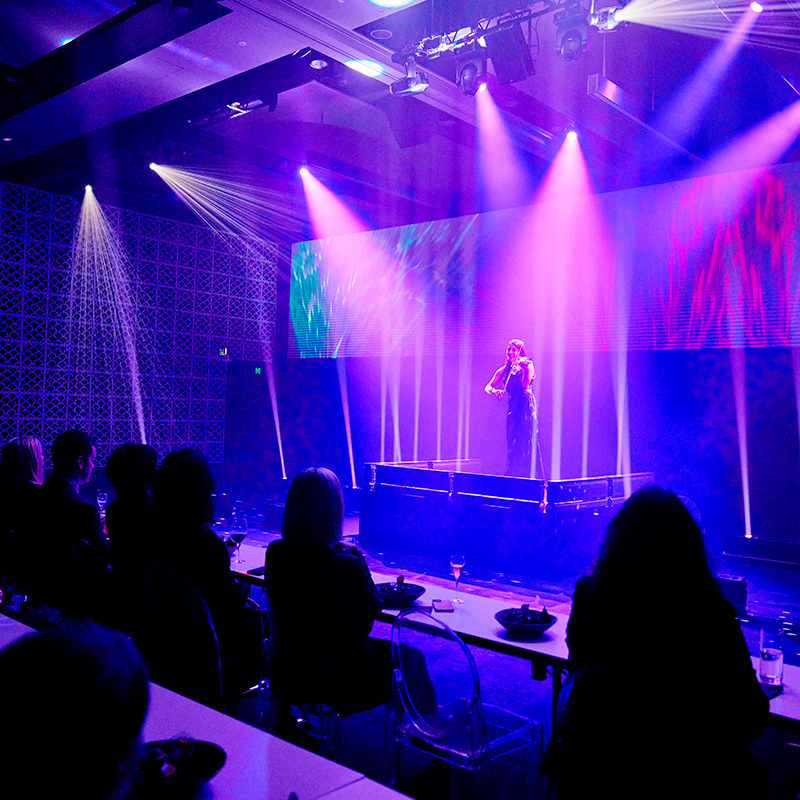 A performer stands on a stage under vibrant purple and pink lights, while an audience seated at tables watches the show in a dark, modern venue—expertly lit by AV company Sydney.