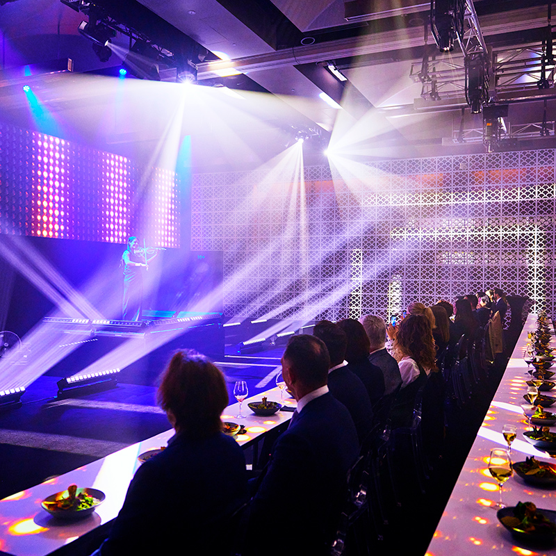 People sit at long tables with food, watching a performer on a lit stage. Bright spotlights and colorful lights illuminate the modern event space, creating a vibrant atmosphere.