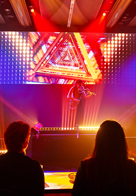 A performer plays an electric violin on stage, illuminated by vibrant red and yellow lights, with a digital triangular tunnel graphic above. As two audience members watch from the foreground, the energy builds in anticipation of an encore.