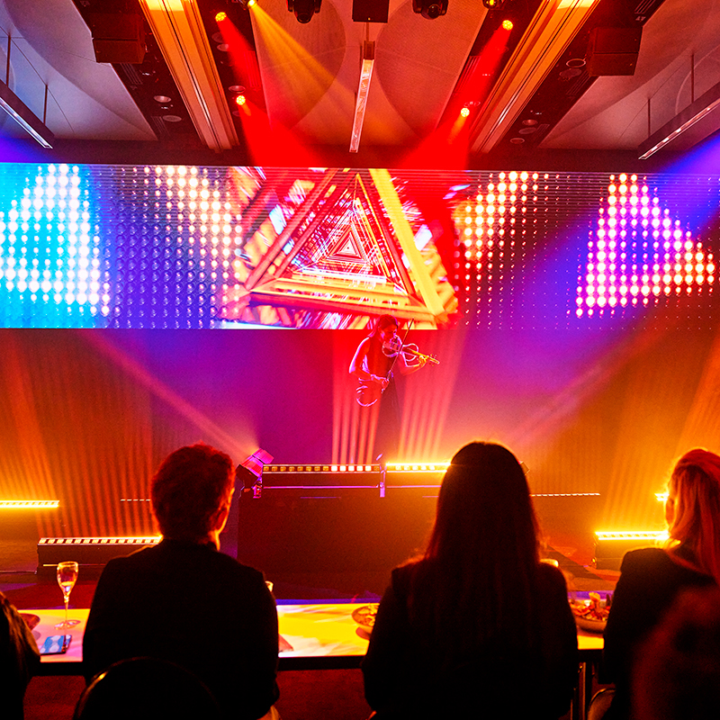 A performer plays the violin on stage with vibrant triangular and LED light displays, while an audience watches from tables in the foreground. The scene is colorful and dynamic, with bright orange and yellow lighting effects.