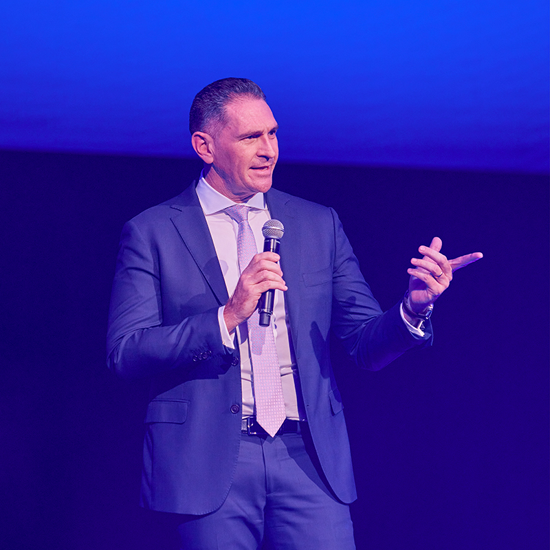 A man in a suit stands on stage holding a microphone, gesturing with his other hand, and speaking under a blue and purple light.