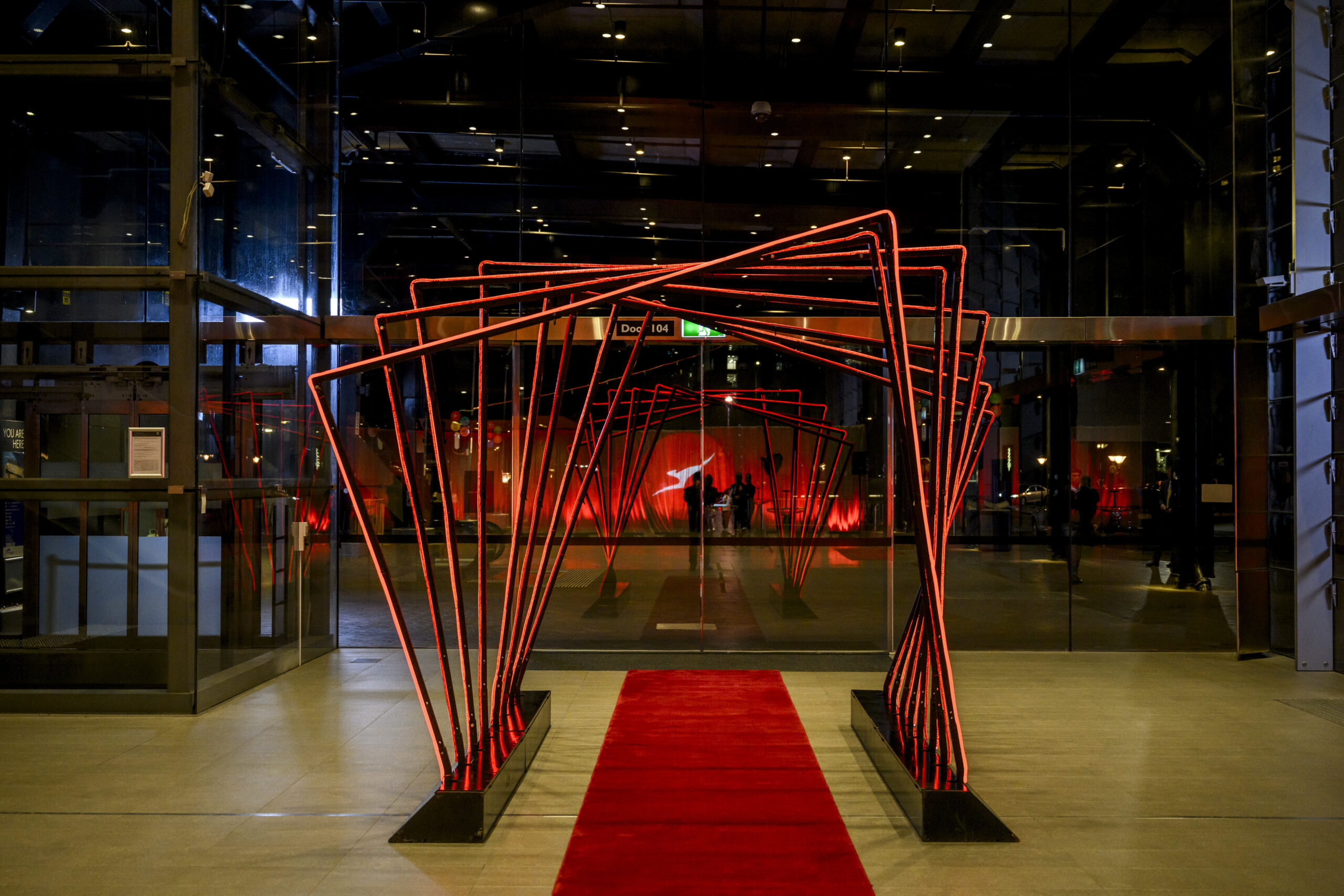 A modern entryway with red metal geometric arches over a red carpet, leading into a building with glass doors and illuminated red lights inside. Reflections and shadows create a dramatic, elegant atmosphere.