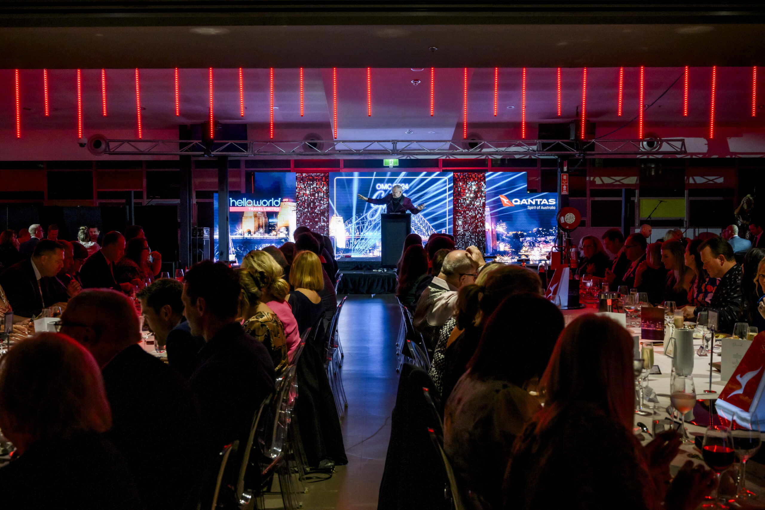 A formal event with guests seated at tables, facing a stage with a speaker. Bright red lights illuminate the ceiling, and digital screens display “helloworld” and “Qantas” logos in the background.