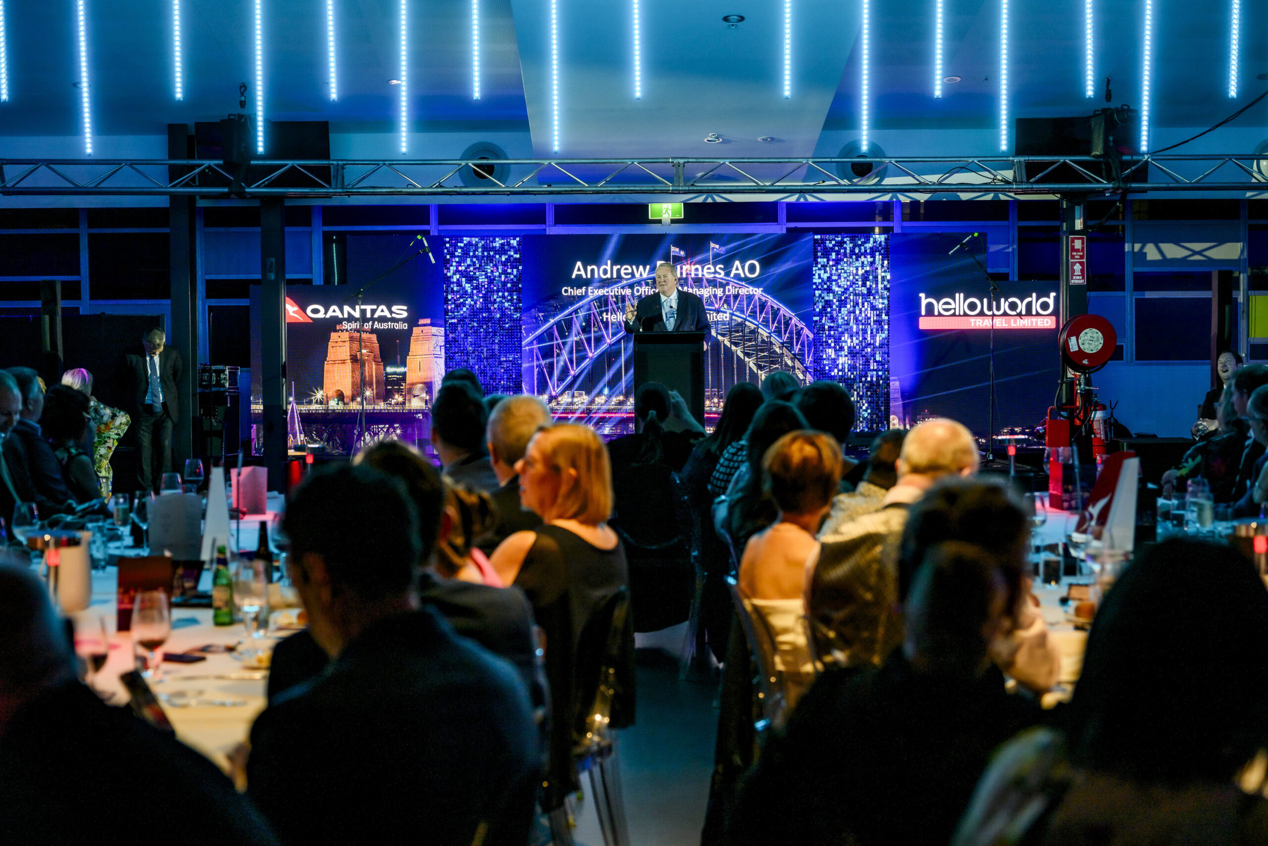 People seated at tables watch a speaker on stage at a formal event with blue lighting. Signs for Qantas and Helloworld are displayed in the background along with an illuminated Sydney Harbour Bridge image.