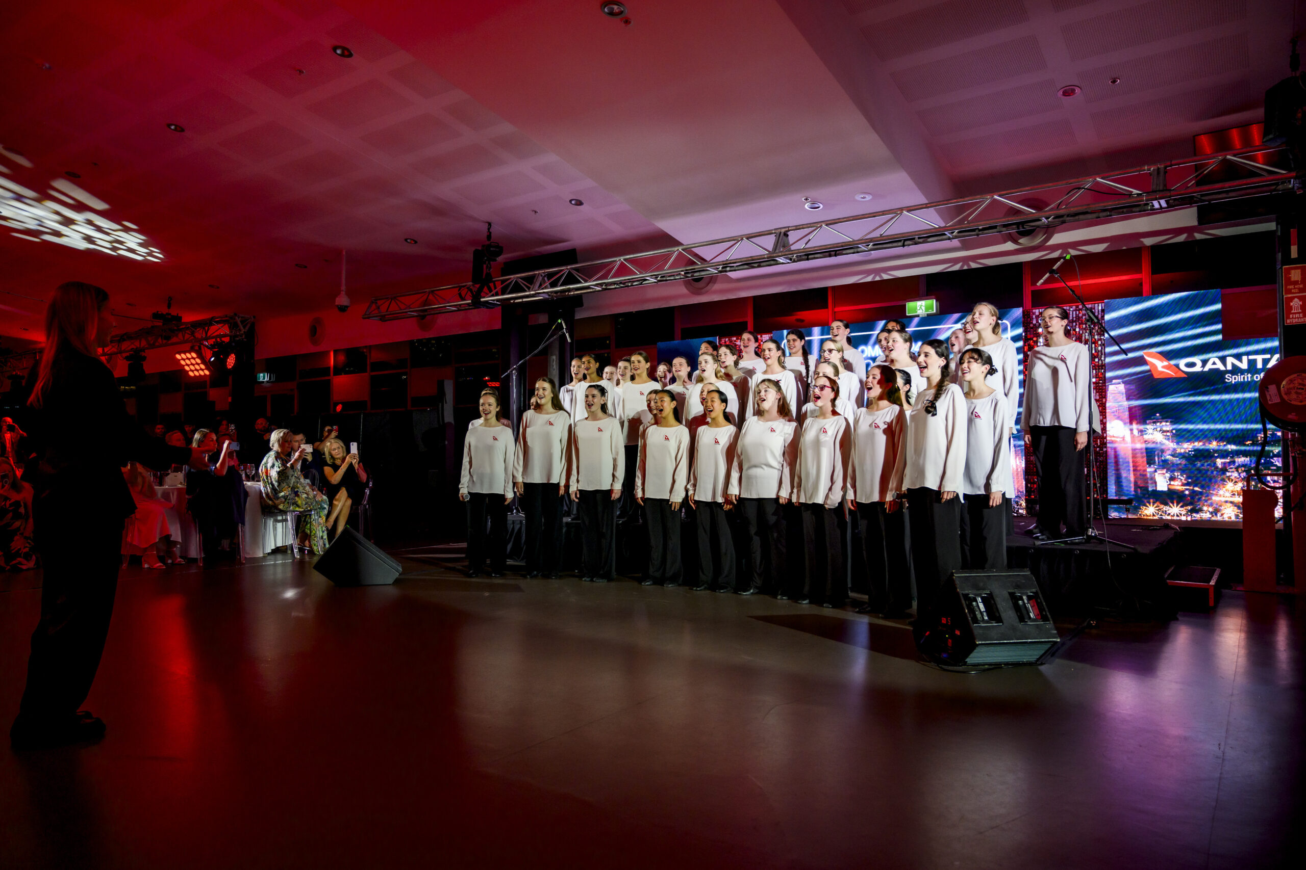 A large choir of people in white tops and black pants sing on a stage under red and purple lighting, with an audience and a large screen in the background.