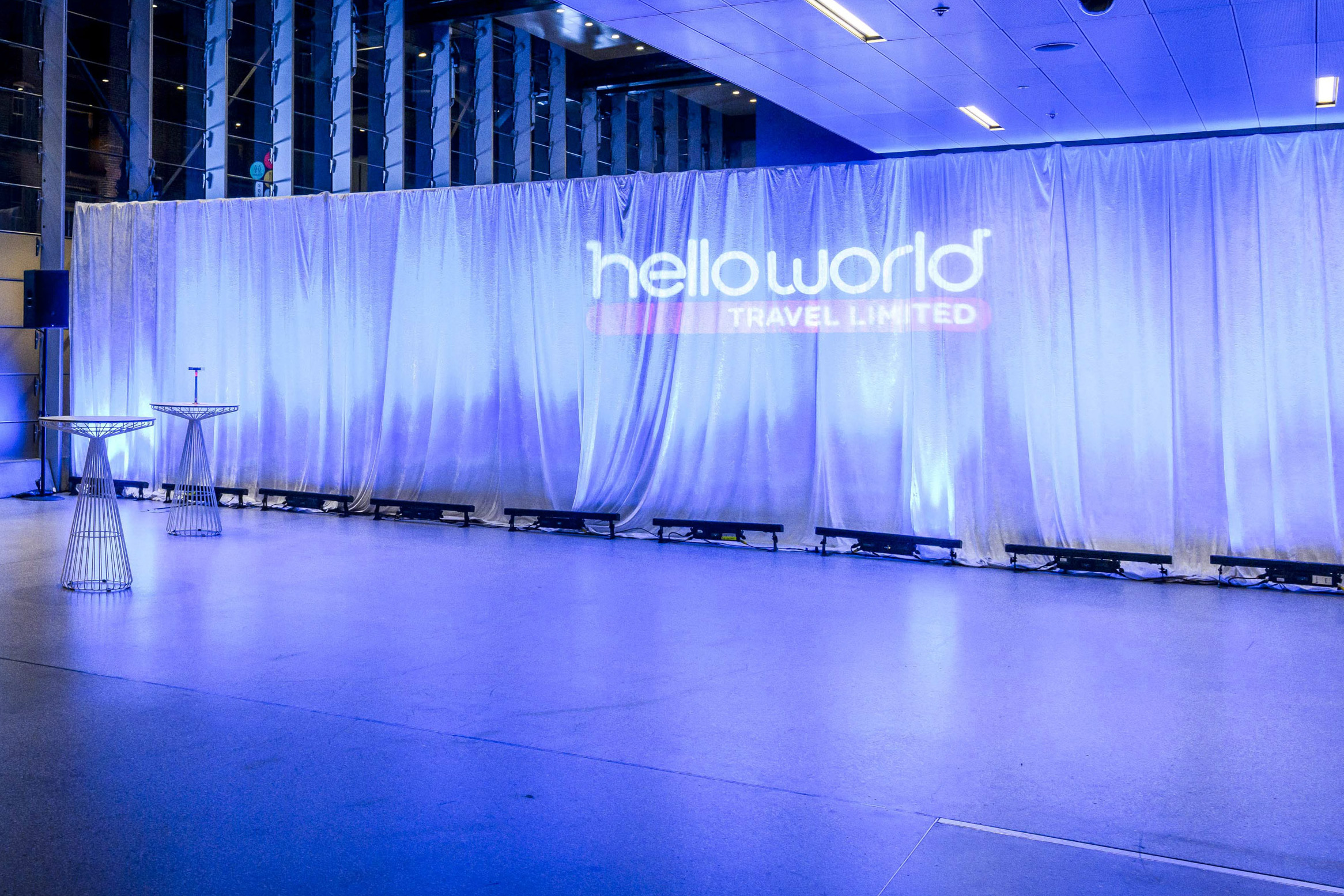 A modern event space with blue lighting, metal cocktail tables, and a large curtain displaying the helloworld TRAVEL LIMITED logo projected on it. The atmosphere is clean and contemporary.
