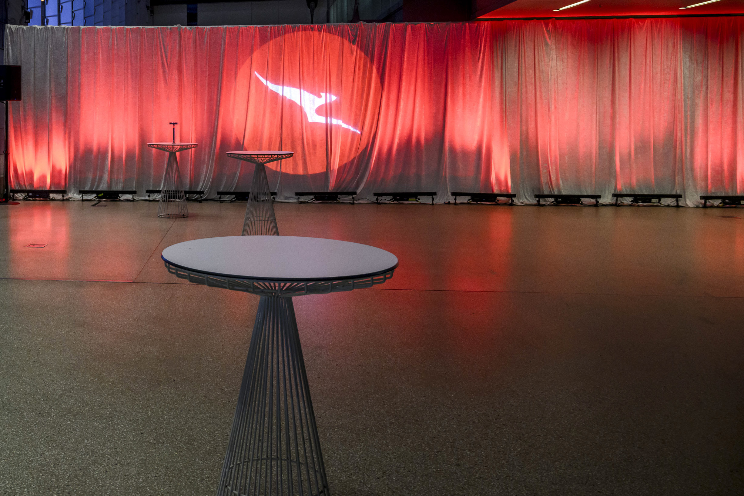 A modern event space with round wire-frame cocktail tables and a red curtain backdrop, featuring a white bird logo projected onto the curtain. The room is empty and well-lit.