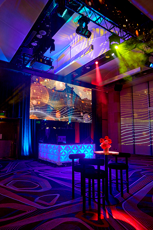 A colorful, modern event space with vibrant lights, a geometric-patterned DJ booth, large digital screens displaying abstract art, and a small table with flowers in the foreground.