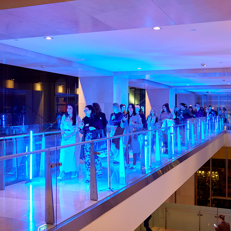 A group of people walk along a modern indoor walkway illuminated with bright blue lights, with glass railings and a reflective floor.