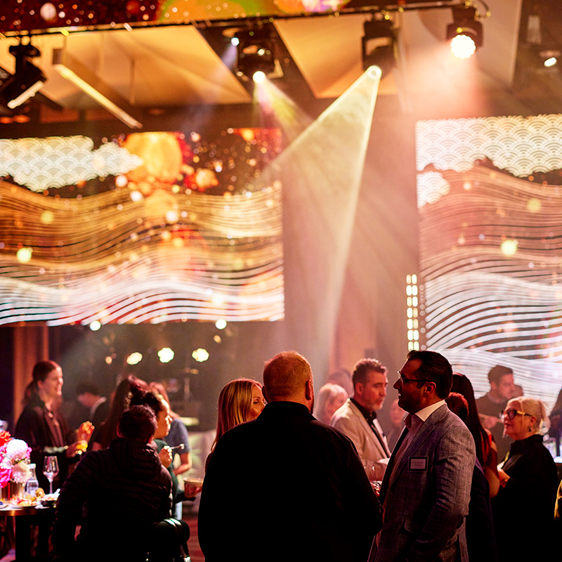 People socialize at a lively indoor event with warm lighting, colorful illuminated screens, and festive decorations in the background. Some guests are seated at tables, while others stand and chat in small groups.