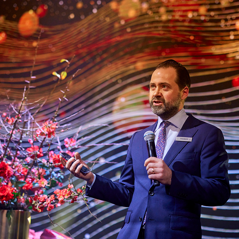 A man in a suit and tie speaks into a microphone while holding a remote, standing next to a floral arrangement. The background features colorful, wavy patterns with orange and yellow lights.