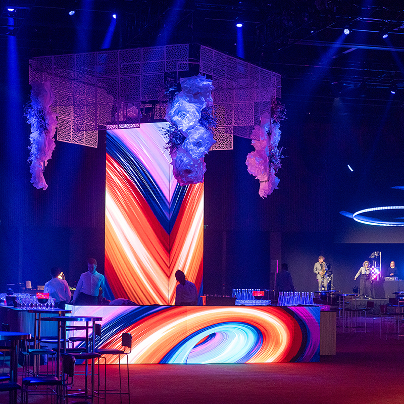 A vibrant, modern event space with colorful swirling LED displays, high tables, and bartenders behind a lit bar. Hanging floral decorations and stage performers are visible under dramatic blue and pink lighting.
