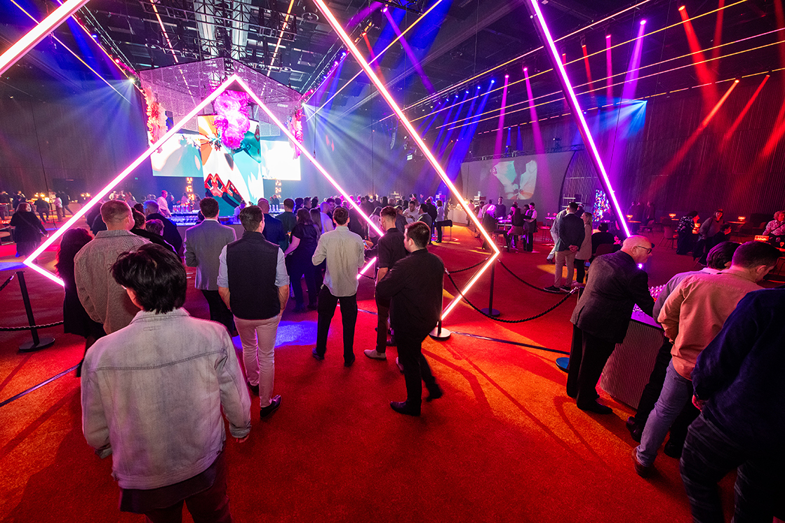 A crowd gathers at an indoor event with vibrant, colorful stage lighting and geometric light structures, creating a lively atmosphere—showcasing the expertise of a leading event production Melbourne company.