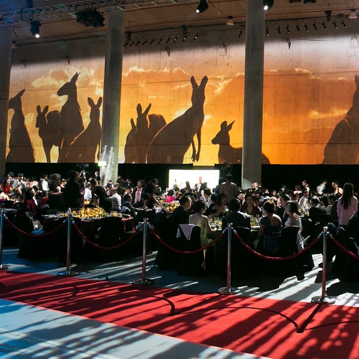 A formal event with guests seated at round tables, red carpet, and a large wall projection of kangaroo silhouettes against a sunset. The venue has tall columns and elegant lighting.