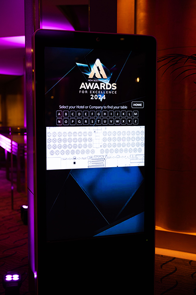 A touchscreen kiosk featuring Event Technology showcases the Awards for Excellence 2024, with an on-screen keyboard for guests to search their hotel or company and find their table, all set against a vibrant purple-lit background.