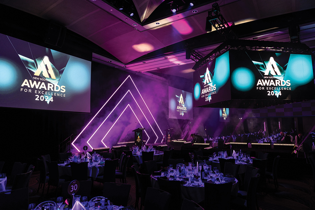 A decorated event hall set up for the 2024 Awards for Excellence, featuring a stage set for gala dinner, illuminated purple lights, round dining tables, and large screens displaying the event name and logo.