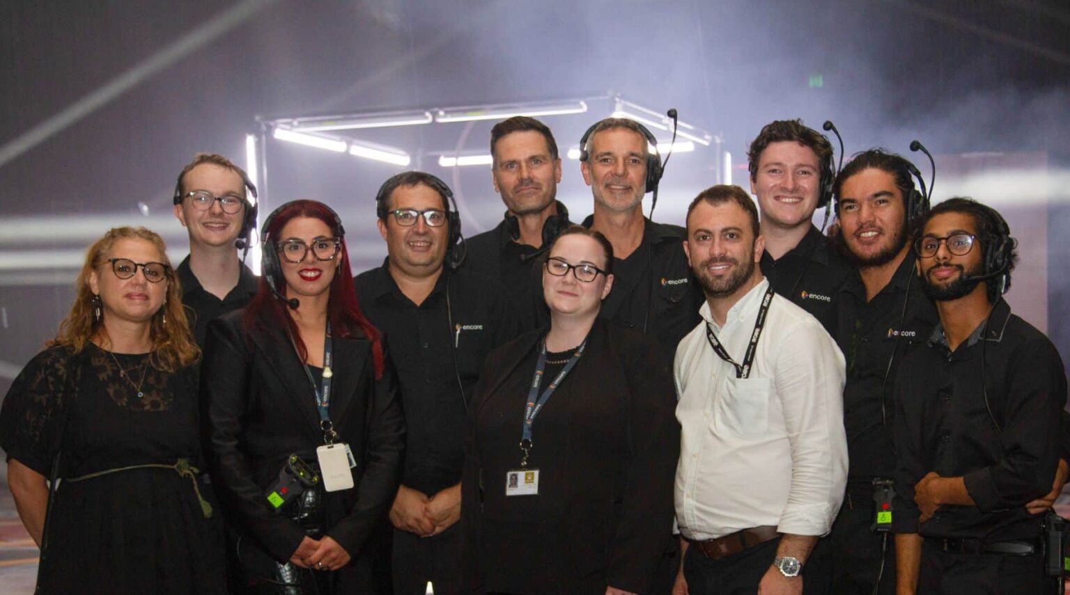 A group of eleven people, some wearing headsets and black outfits, pose together indoors in front of a misty, illuminated background, smiling at the camera.