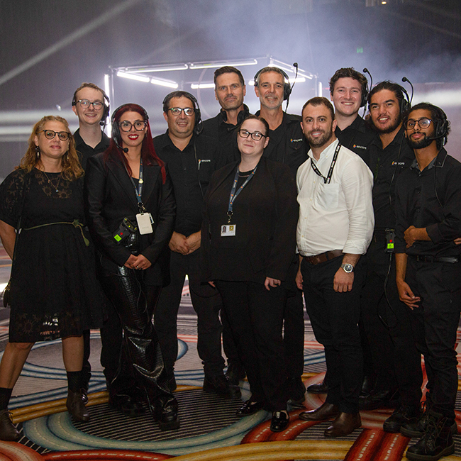 A group of eleven people, dressed mostly in black, pose together indoors on a patterned floor with stage lighting and fog in the background. Some wear headsets and lanyards, suggesting an event or production team.